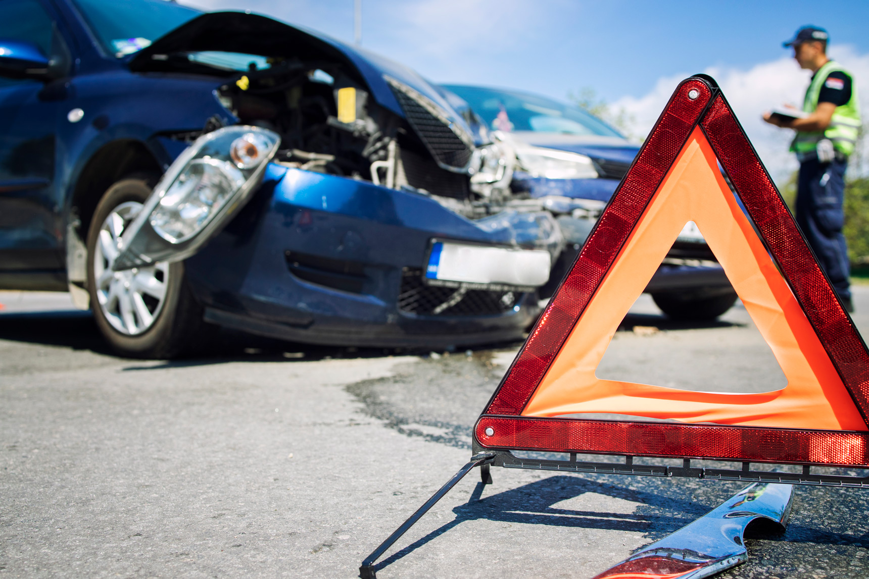  An image of a car crash with a warning triangle in the foreground and a police officer in the background.